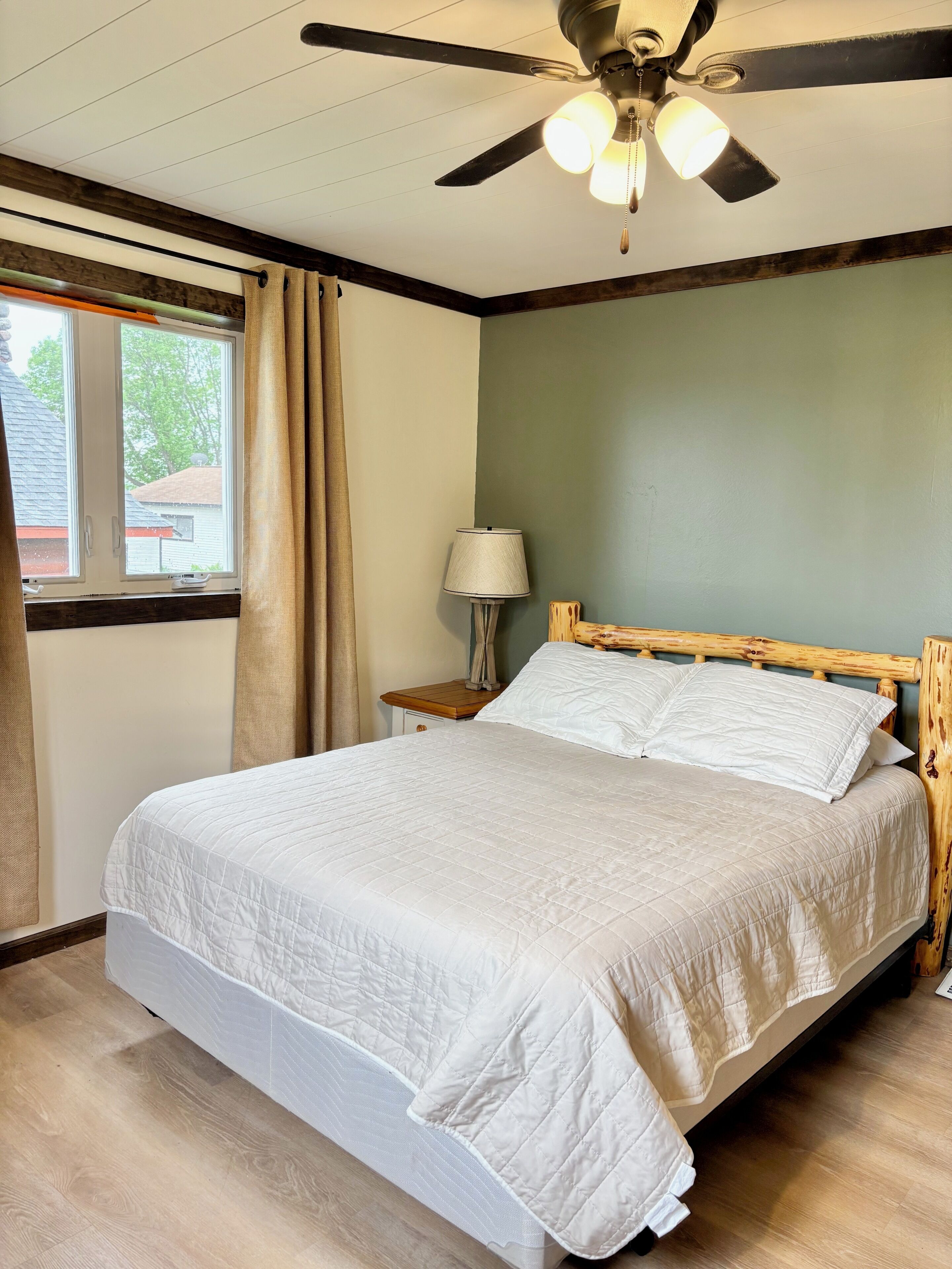 A bedroom with a bed, a ceiling fan, a window with curtains, and a nightstand with a lamp.