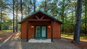 Una cabaña de troncos con puerta verde y dos ventanas, rodeada de árboles.