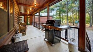 Un porche cubierto con una parrilla y zona de estar, una terraza de madera y vistas al bosque circundante.