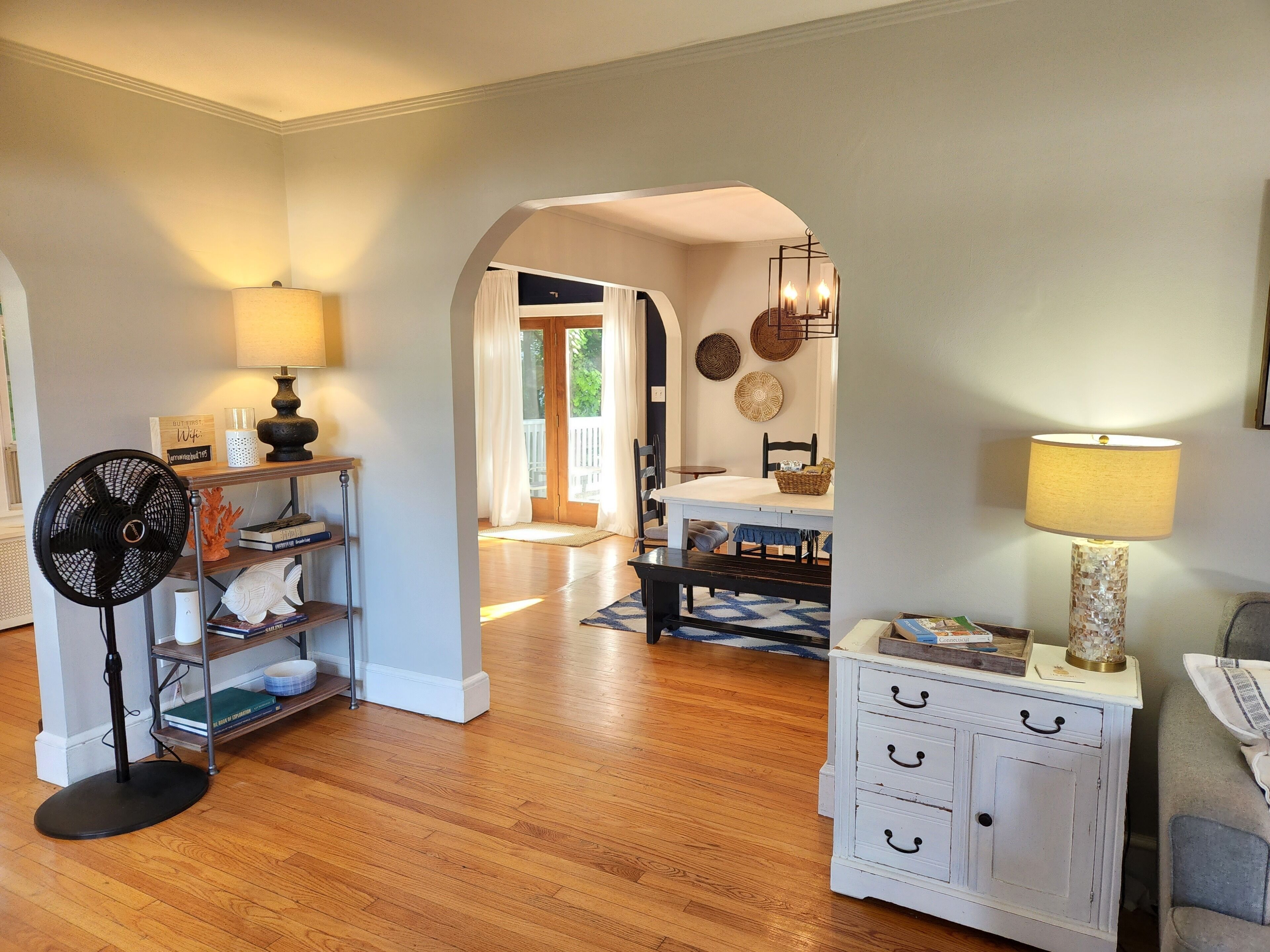 Une pièce avec un plancher en bois, une commode blanche à tiroirs, un ventilateur, une table avec une lampe et une vue sur un balcon, visible à travers une arche.