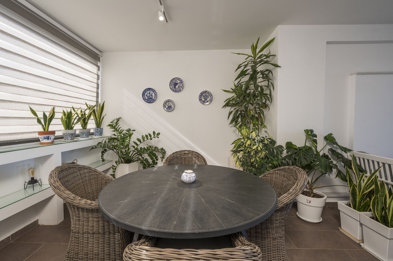 A round table with wicker chairs, surrounded by potted plants and decorative plates on the wall.