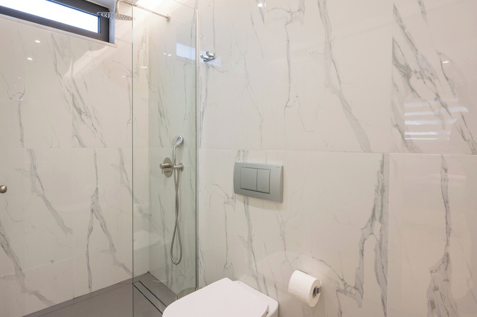 Et moderne badeværelse med et glasafskærmet brusebad, marmorvægge, toilet og en håndtørrer.