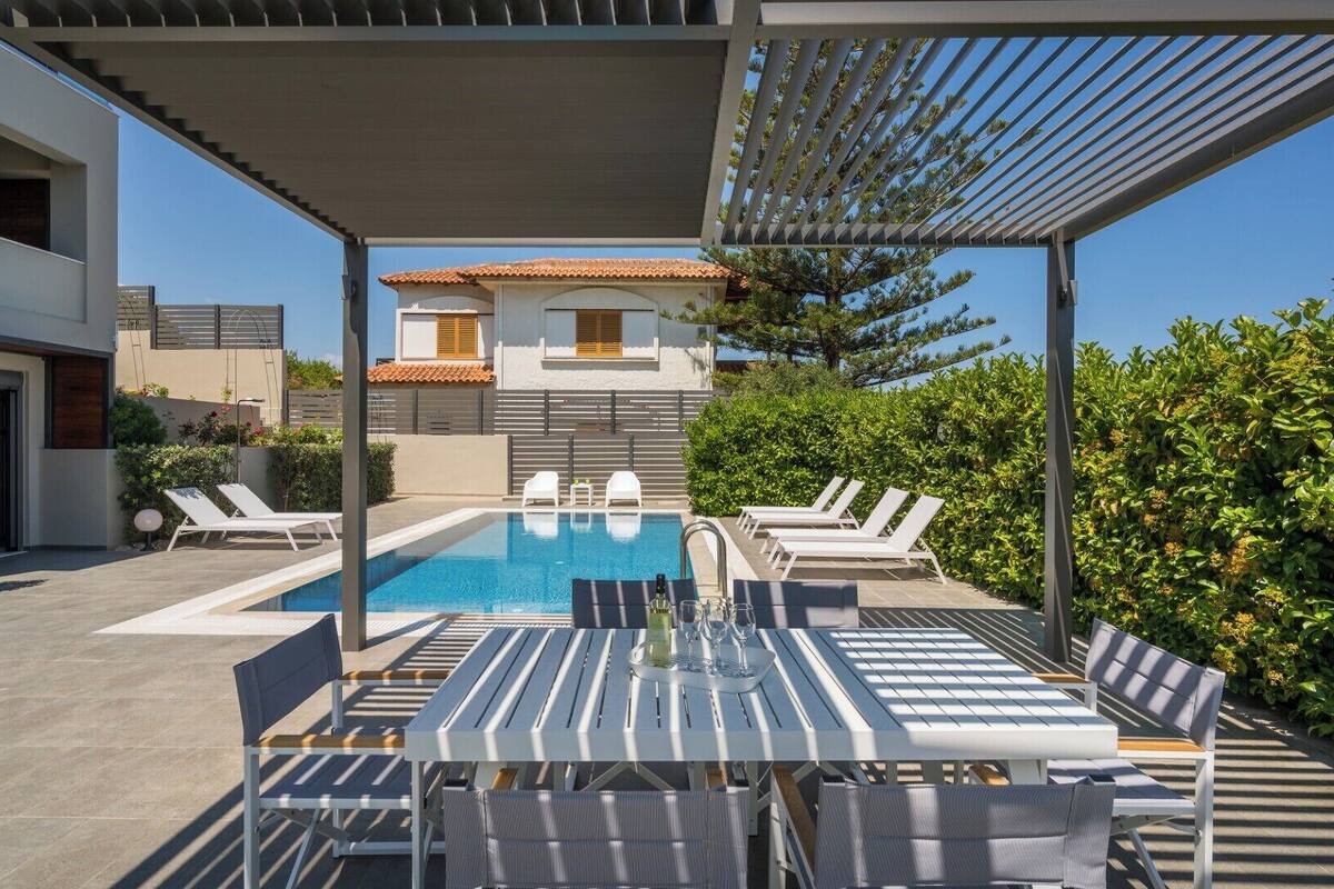Un espace piscine extérieur moderne, doté de chaises longues et d’une table, entouré de verdure et bordé d’un bâtiment dont le toit est recouvert de tuiles.