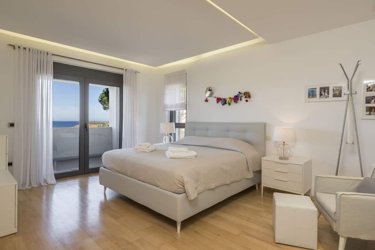 Une chambre moderne avec un grand lit, une table de chevet, une chaise et une vue sur la mer, accessible par une porte coulissante.