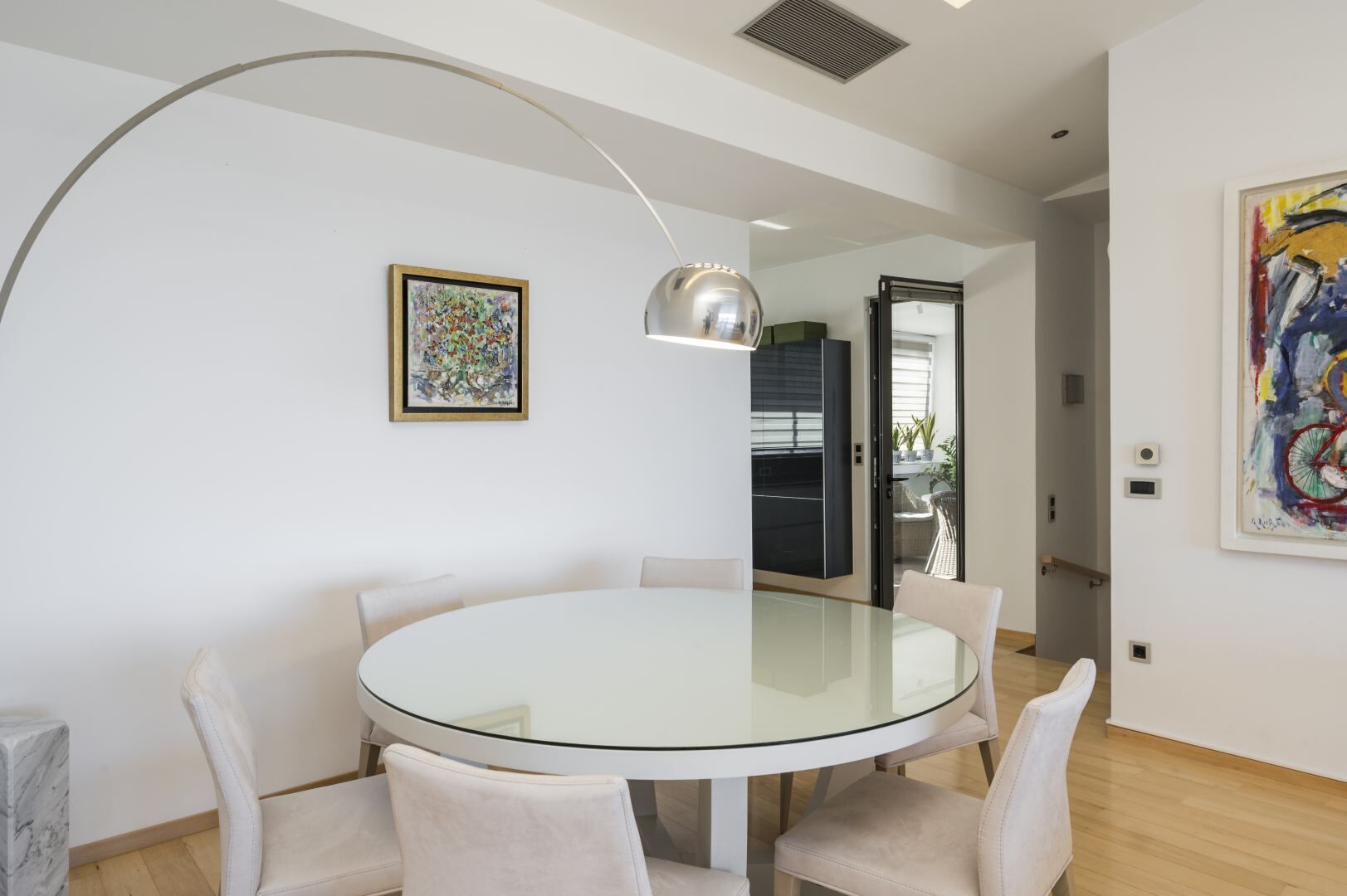 A modern dining area with a glass table and white chairs, a pendant light, and two framed artworks on the walls.