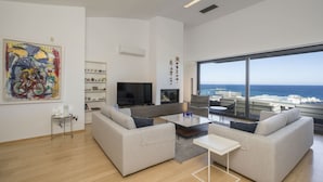 Een moderne woonkamer met een groot raam met uitzicht op de oceaan, een flatscreen televisie en een boekenkast.
