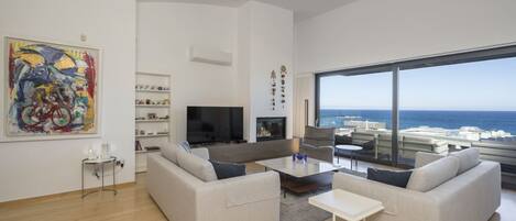 A modern living room with a large window overlooking the ocean, a flat-screen TV, and a bookshelf.
