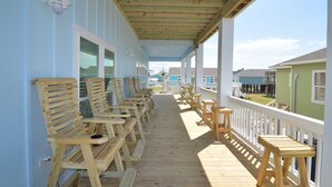 A covered porch with wooden chairs and tables, a white railing, and a view of neighboring houses.