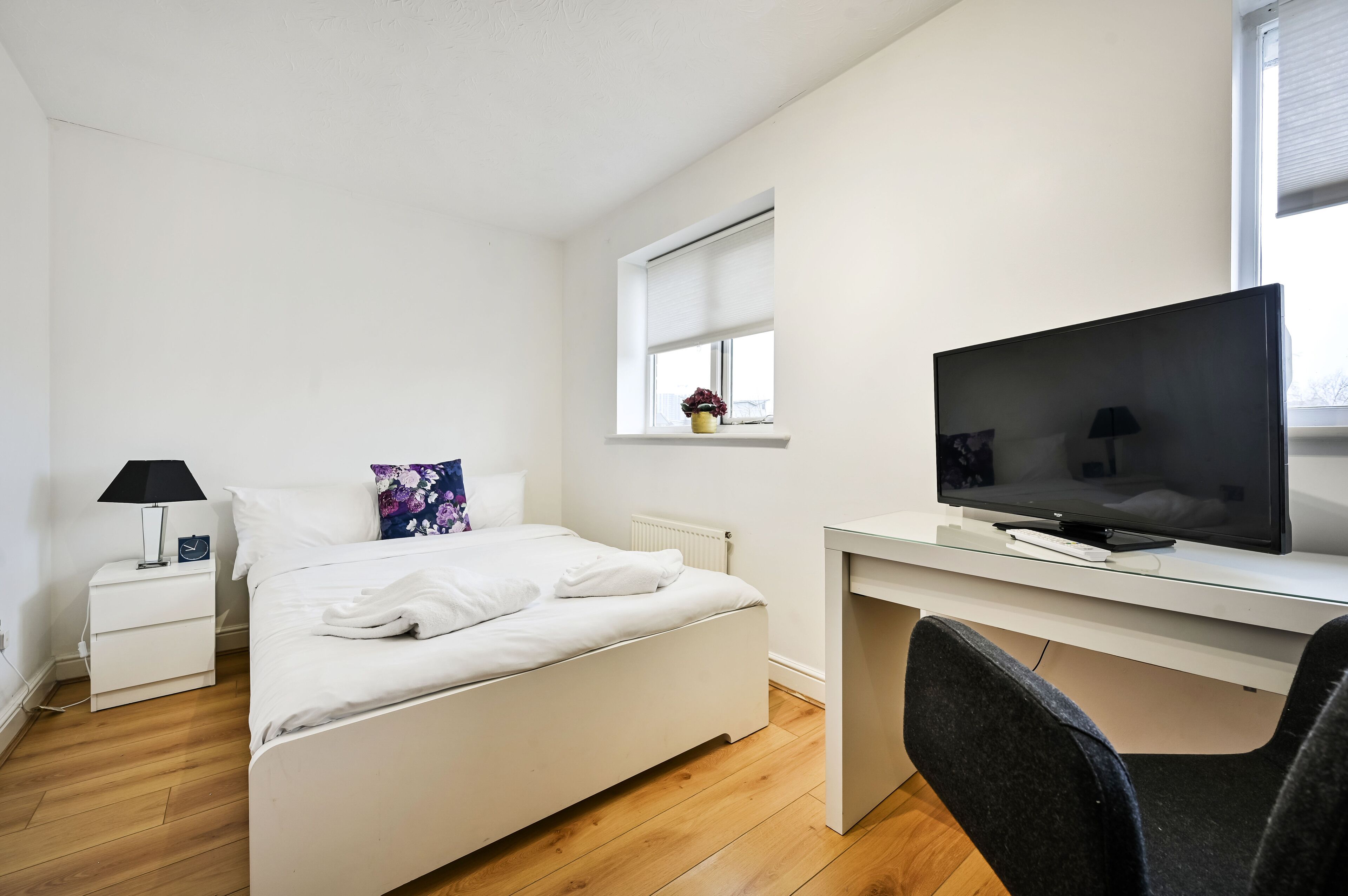 A bedroom with a bed, a desk, a television, a chair, a nightstand, a lamp, and a window.
