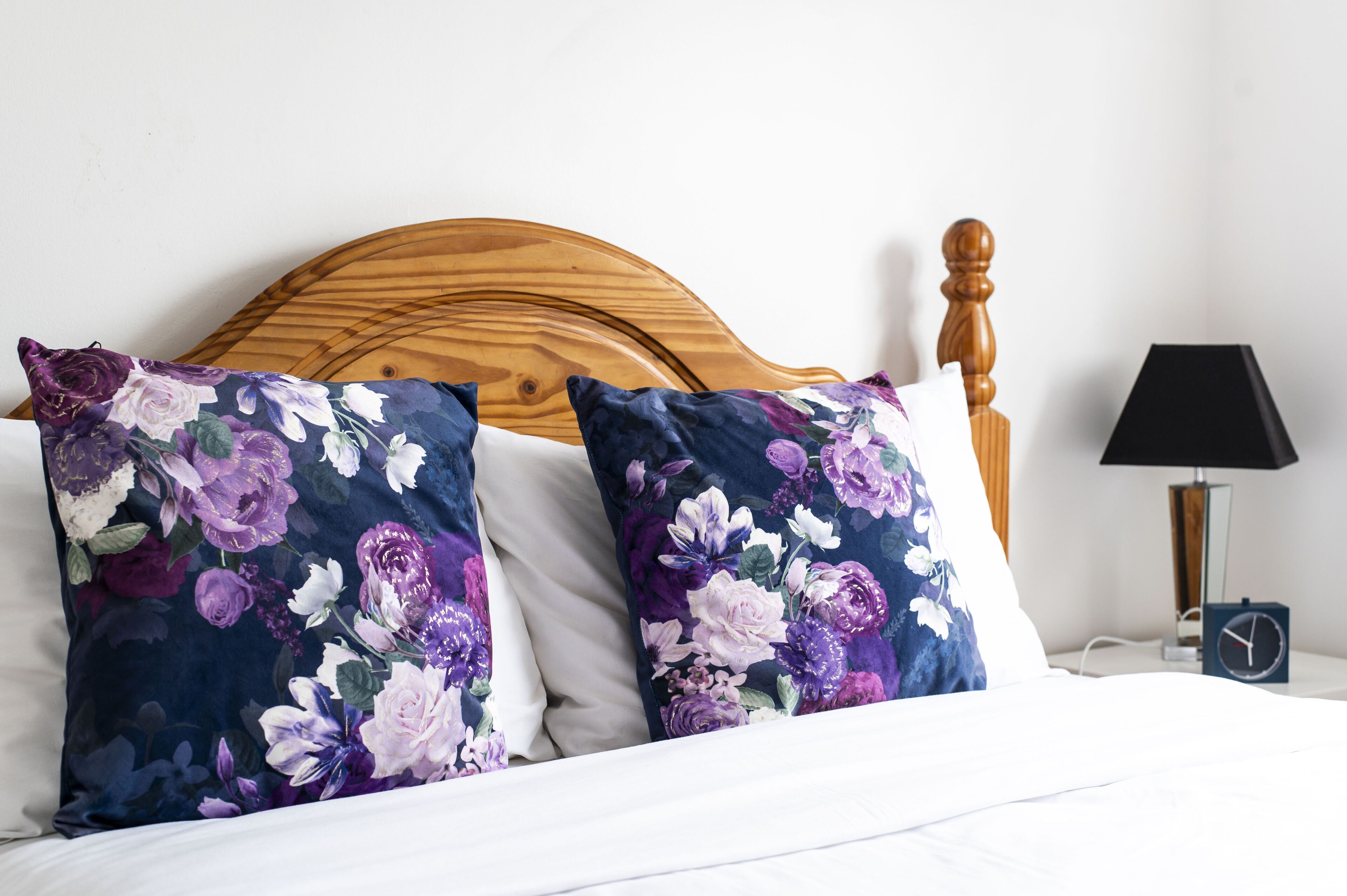 A bed with floral pillows, a wooden headboard, a bedside table with a lamp, and a clock.