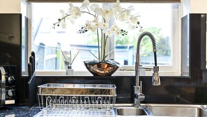 A kitchen with a black countertop, a stainless steel sink, a dish rack, and a vase with white flowers on the windowsill. - Riverside Townhouse with private Garden (London)