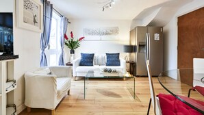 A modern living room with a white sofa, a glass coffee table, and a red chair. There is a large window with blue curtains, a painting on the wall, and a refrigerator. - Riverside Townhouse with private Garden (London)