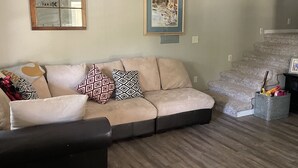 Sala de estar com sofá seccional, ventilador de teto, quadro emoldurado, janela refletindo luz e escada.
