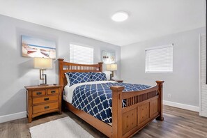 Une chambre à coucher avec un lit en bois, deux tables de chevet, une commode, un tapis blanc et un luminaire au plafond.