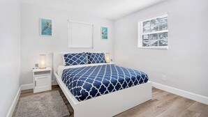 Um quarto com cama branca, roupa de cama estampada em azul, mesinha de cabeceira pequena com abajur e uma janela com vista para a cidade.