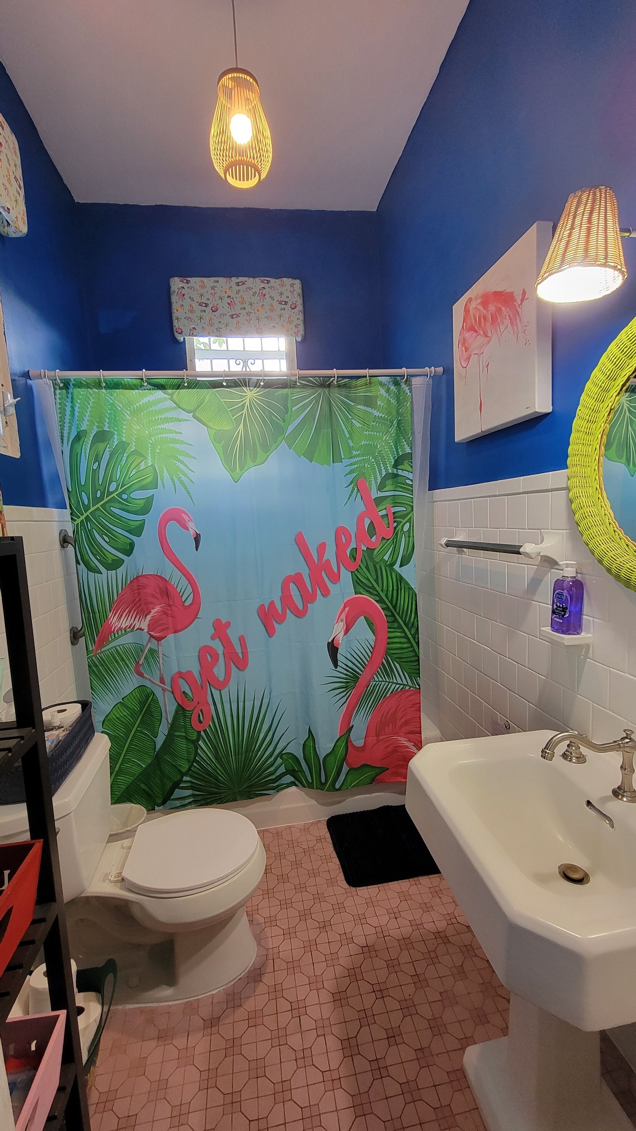 A bathroom with a blue wall, a shower curtain with flamingos and tropical leaves, a toilet, a sink, and a mirror.