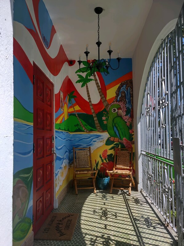 Un pasillo con un mural colorido, una puerta roja, dos sillas de mimbre, un portón metálico y un suelo estampado.