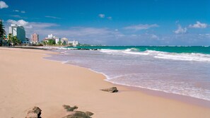 Una playa con agua turquesa cristalina, una orilla arenosa y edificios al fondo.