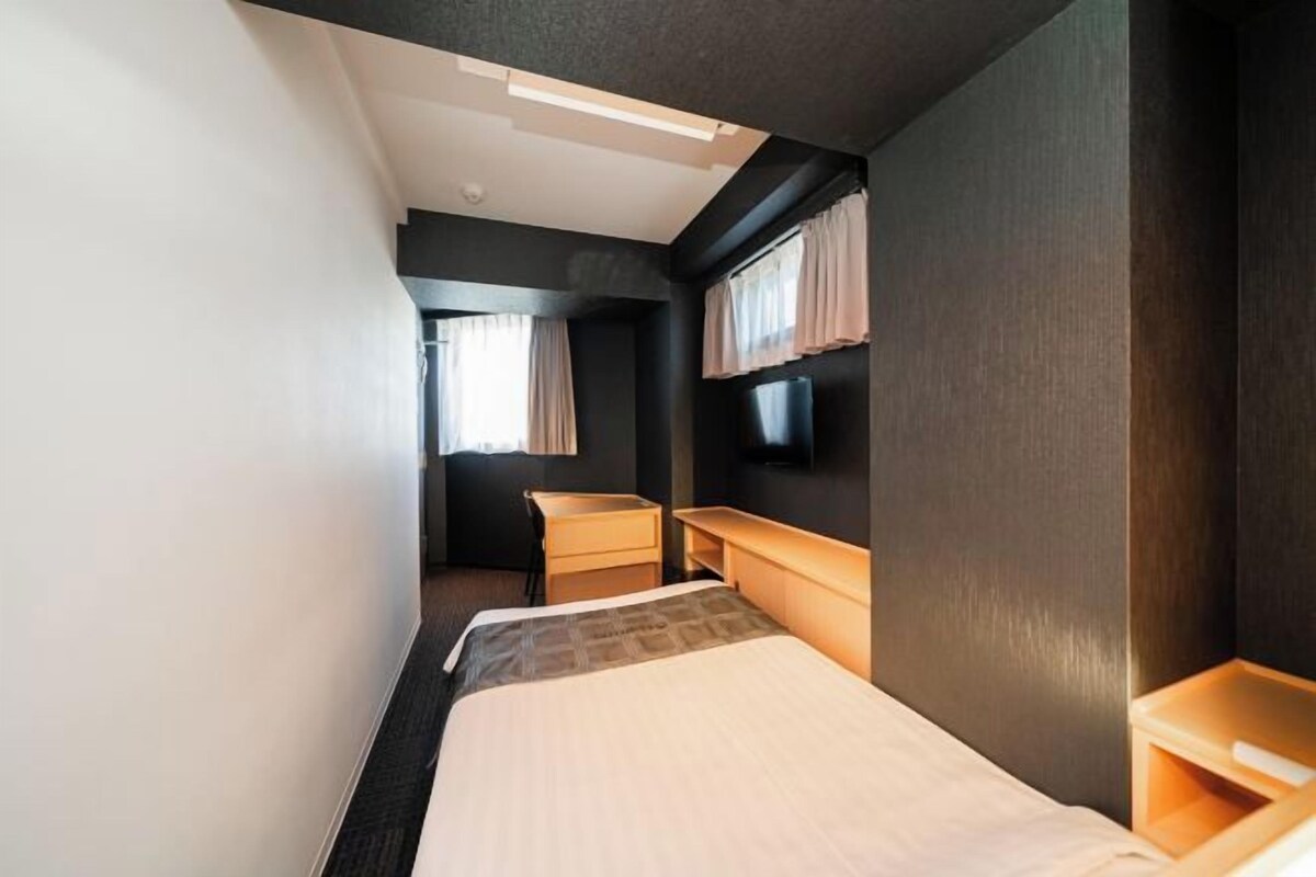 A compact hotel room with a bed, bedside table, a small desk, a flat-screen TV mounted on the wall, and a window with curtains.