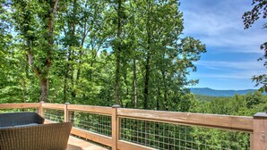 Une terrasse en bois avec un fauteuil en osier, entourée d’une rambarde en métal, donnant sur une zone boisée avec des montagnes à l’horizon.