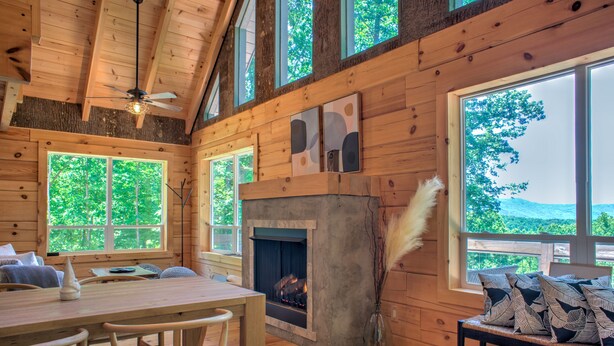 L’intérieur d’un chalet en bois comprend un coin repas, une cheminée et de grandes fenêtres donnant sur un paysage verdoyant.