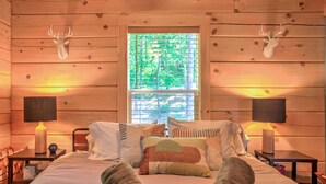 Une pièce de style cabane en bois, comprenant un lit, deux lampes fixées au mur, une fenêtre avec des stores et deux décorations murales représentant des têtes de cerf.