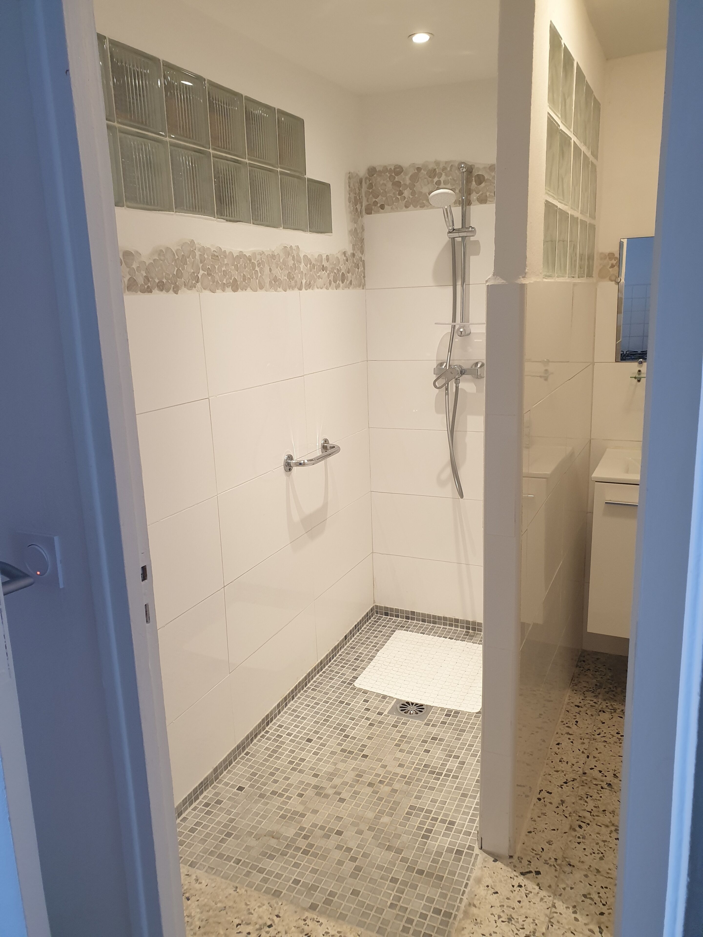 Un espace douche doté d’une porte en verre, d’un mur recouvert de carreaux blancs et d’un petit tapis blanc posé sur le sol.