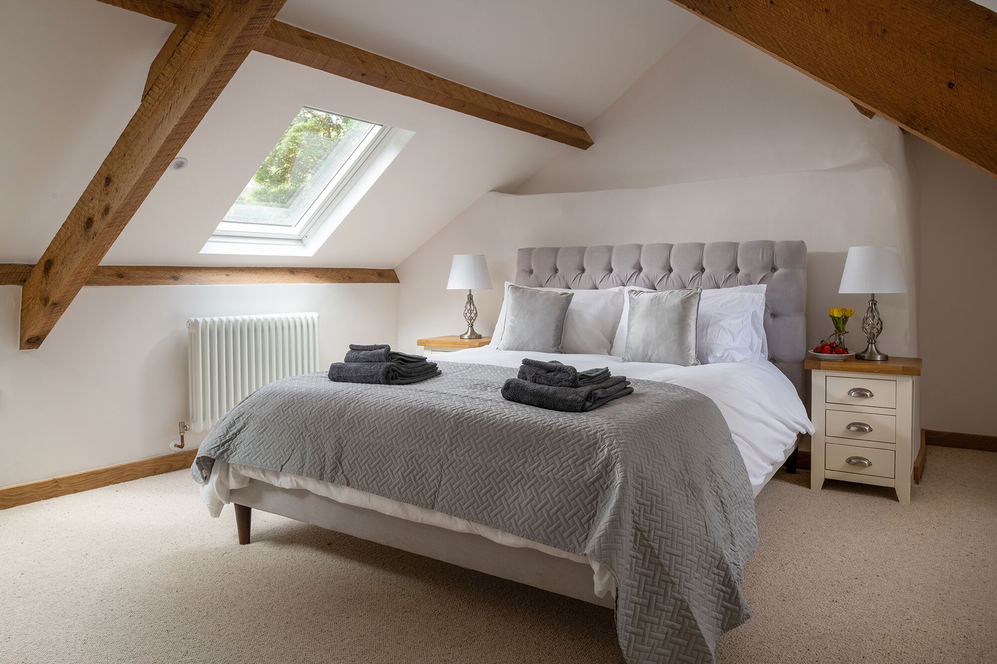 A bedroom with a sloped ceiling, a bed with a grey quilt, two bedside lamps, and a view of greenery through a skylight.