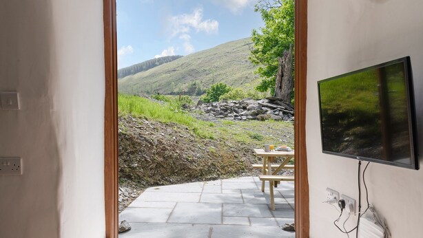 A doorway leading to an outdoor patio with a table and chairs, a television mounted on the wall, and a view of a hillside. - Snowdonia Cottage with Stunning views of Bala lake and mountains (Llangower)