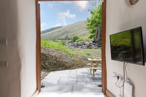 Una puerta que da a un patio al aire libre con una mesa y sillas, un televisor montado en la pared y vistas a una colina.