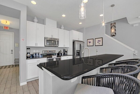 A modern kitchen with white cabinets, a black countertop island, stainless steel appliances, and wicker chairs.