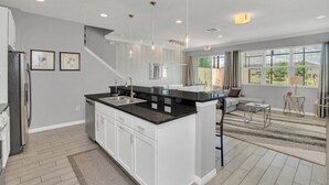 A modern kitchen with white cabinets, black countertops, and stainless steel appliances. There is a large island with a sink and a dining area with a sofa and a small table. - Beautiful TownHome With Pool Near Disney! (Kissimmee)