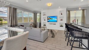 A modern living room with a large TV, a dining area with black bar stools, and a view of a lake. - Beautiful TownHome With Pool Near Disney! (Kissimmee)