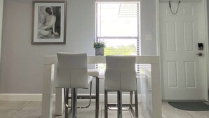 A dining area with two white chairs and a table, a framed artwork on the wall, a window with blinds, and a white door with a chain. - Welcome to this stylish and spacious luxury suite (Fort Lauderdale)