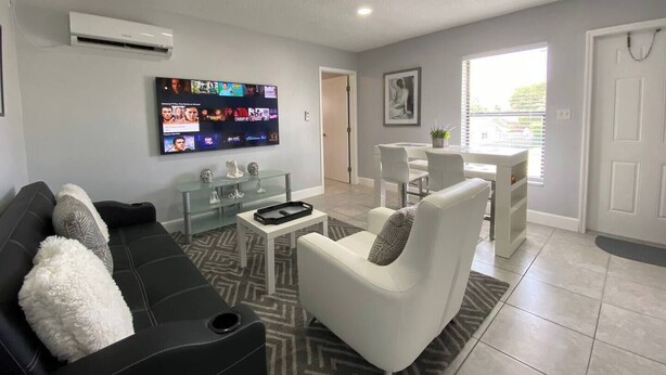 A modern living room with a black sofa, white armchair, and a glass coffee table. A flat-screen TV is mounted on the wall, and there is a small dining area with a white table and chairs. - Welcome to this stylish and spacious luxury suite (Fort Lauderdale)