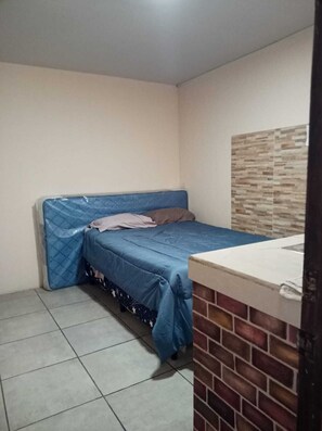 Ein kleines, schlichtes Zimmer mit einem Einzelbett, Fliesenboden und einer Wand mit Ziegeloptik.