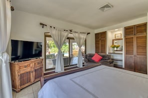 Une chambre avec un grand lit, un téléviseur posé sur un meuble en bois, un canapé agrémenté d’un coussin rouge et une porte-fenêtre coulissante donnant sur un espace extérieur.