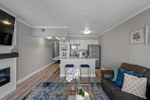 A modern living room with a fireplace, a sofa with cushions, a coffee table with a plant, and a kitchen in the background.