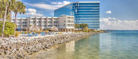 Ein Strandhotel mit einem modernen Glasgebäude, felsigem Ufer, klarem Wasser und Palmen.