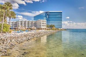 A beachfront hotel with a modern glass building, a rocky shoreline, clear water, and palm trees.