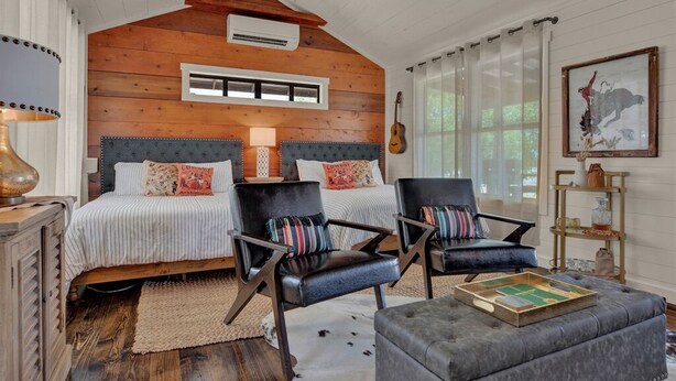 A cozy bedroom with a wooden ceiling, a large bed with striped bedding, two black leather chairs, a small ottoman, and a guitar on the wall. - Cozy Chic Cabin on Texas Wine Trail (Stonewall)