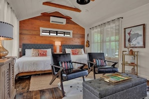 A cozy bedroom with a wooden ceiling, a large bed with striped bedding, two black leather chairs, a small ottoman, and a guitar on the wall.