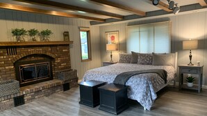Un dormitorio con chimenea de ladrillo, cama con ropa de cama estampada, mesita de noche con lámpara y ventana con persianas.
