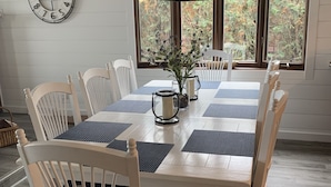 Un comedor con una mesa y sillas blancas, una lámpara colgante, un reloj en la pared y una ventana con vistas a árboles.