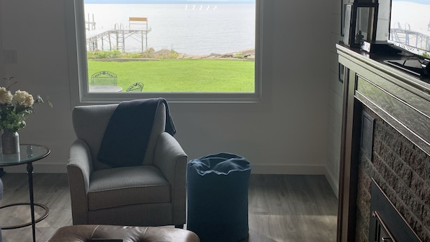 Una habitación con un ventanal grande que da a un área verde y una masa de agua. Hay una silla, un sillón de peluche azul y una pequeña mesa con un jarrón de flores.