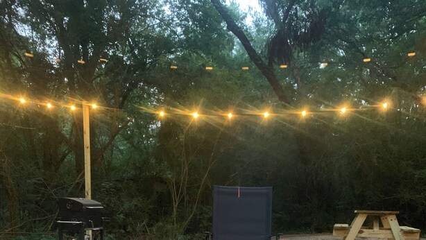 A campfire surrounded by rocks, a wooden bench, and a picnic table in a wooded area with string lights. - Minutes from Beautiful broken bow lake  (Broken Bow)