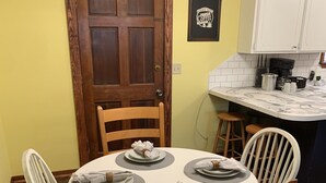 A dining area with a round table set for four, surrounded by wooden chairs. A wooden door and a kitchen counter with a coffee maker are visible in the background. - Minutes from Beautiful broken bow lake  (Broken Bow)
