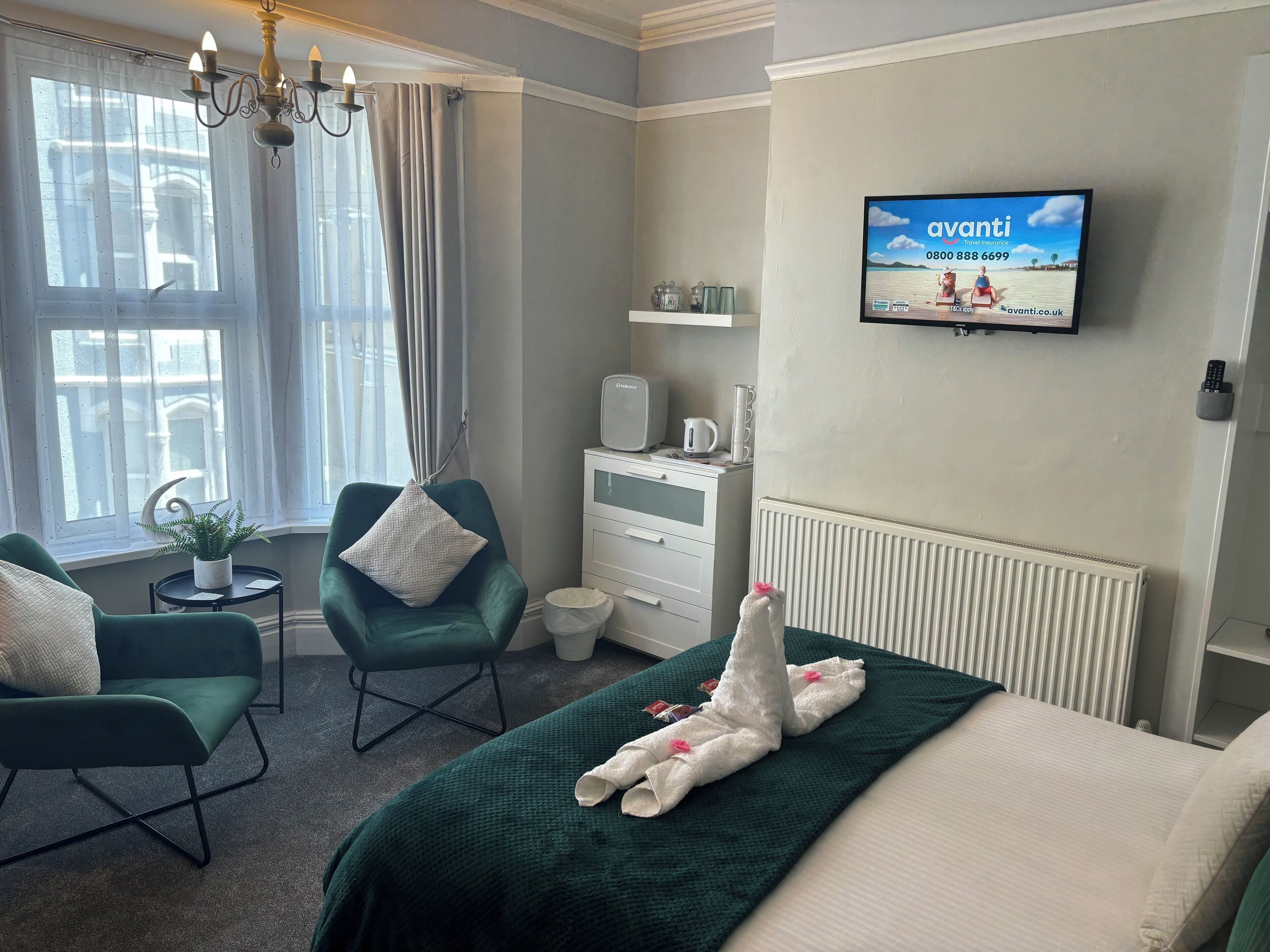 A hotel room with a bed, two green armchairs, a small table with a plant, a TV displaying an advertisement, and a radiator.