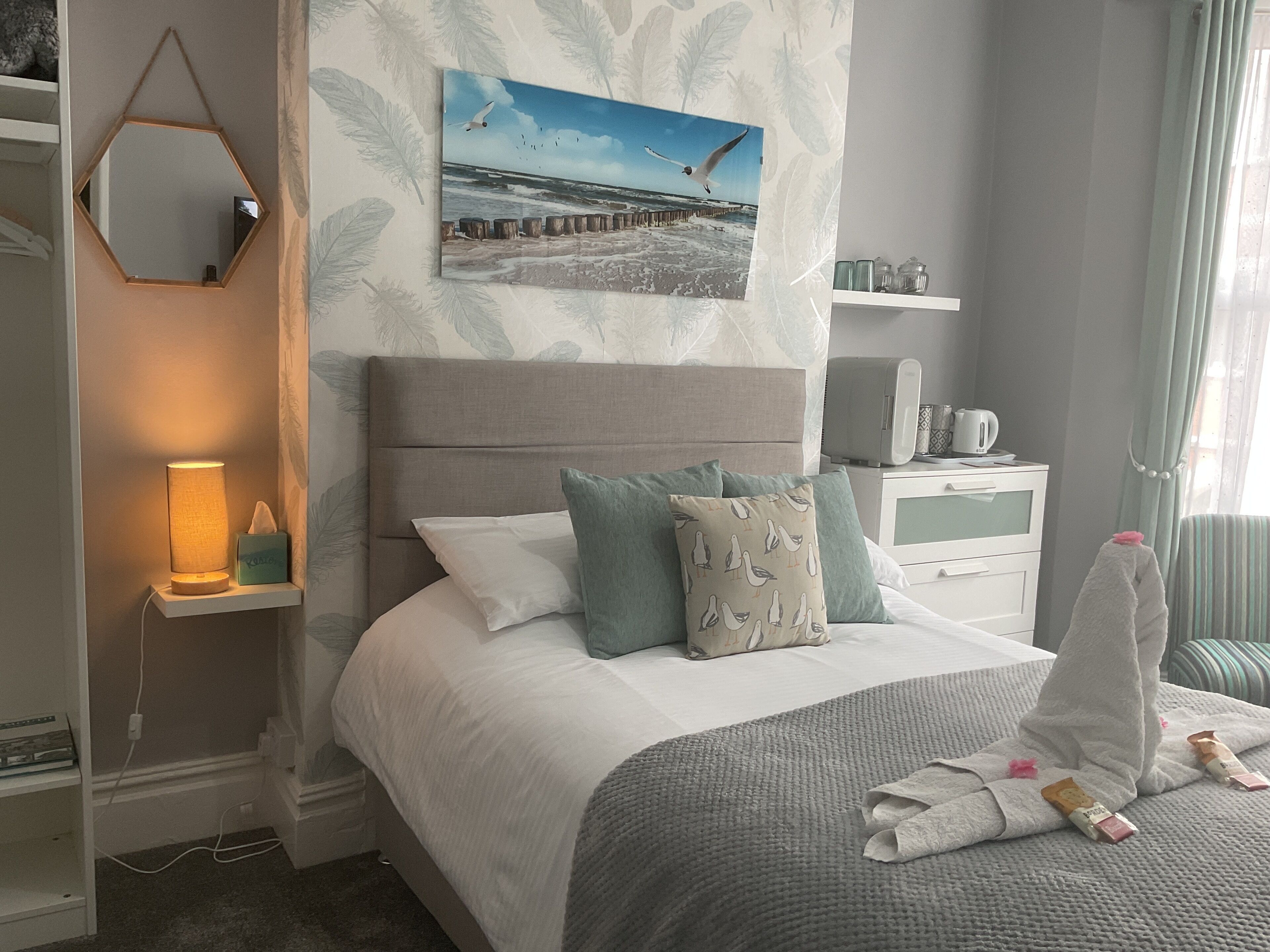 A bedroom with a bed, pillows, a bedside table with a lamp, a nightstand with a kettle, and a wall with a seascape painting.
