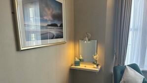 Una camera d'albergo con un quadro paesaggistico incorniciato, un comodino con una lampada, uno specchio e una poltrona verde con un cuscino.